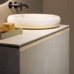 Modern bathroom vanity with a round marble vessel sink and wall-mounted faucet; on the counter is a tray with soap and a sponge/bread-like item.