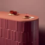 Two amber glass mugs with red tea sit on a round wooden tray with a cookie, plus a second mug on a napkin on a pink curved counter against a pink wall.